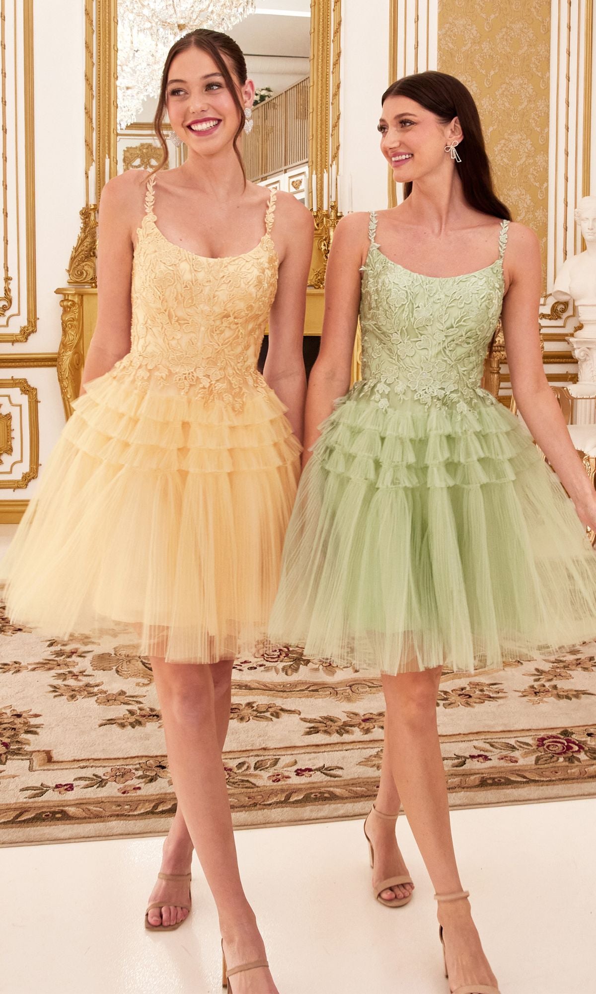 Two women stand smiling in an ornate room, each wearing the Lace-Bodice Tiered-Skirt Short Prom Dress 9310—one in pale yellow, the other in light green—both featuring lace bodices and layered tulle skirts for a charming party look.